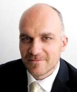 Close-up portrait of a bald man in a suit and tie against a white background.