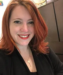 Smiling woman with shoulder-length red hair wearing a black top and gold necklace.