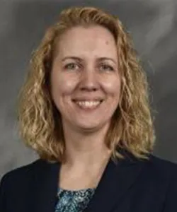 Smiling woman with curly blonde hair wearing a dark blazer and patterned blouse against a gray background.