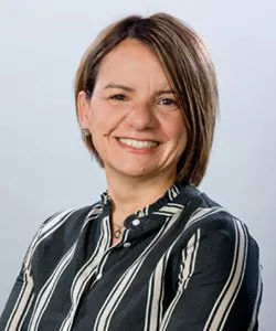 Smiling woman with short brown hair wearing a black and white striped blouse against a light gray background.