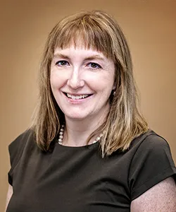 Smiling woman with shoulder-length blonde hair wearing a black top and a pearl necklace against a neutral background.
