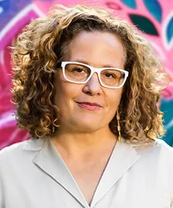 Woman with curly hair and white glasses wearing a light-colored collared shirt, posed against a colorful background.