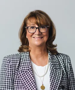 Smiling woman with brown hair wearing glasses, a black and white checkered blazer, white top, and a necklace with a round pendant.