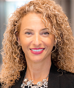 Smiling woman with curly blonde hair wearing a black blazer and floral necklace.