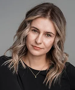 Woman with shoulder-length blonde hair and soft waves wearing a black top and a delicate necklace against a gray background.