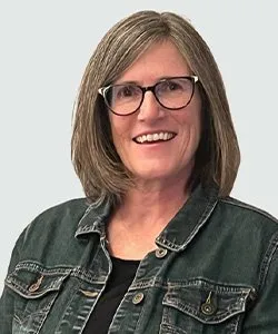 Smiling woman with shoulder-length brown hair and glasses wearing a dark denim jacket over a black top against a light gray background.