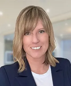 Smiling woman with shoulder-length blonde hair wearing a navy blazer and white shirt in a bright indoor setting.