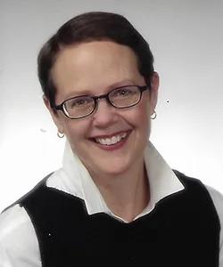 Smiling woman wearing glasses, a white collared shirt, and a black vest against a plain white background.