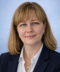 Portrait of a woman with shoulder-length light brown hair, wearing a navy blazer, white blouse, small hoop earrings, and a pendant necklace, smiling softly against a light blue background.