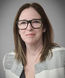 Middle-aged woman with straight brown hair wearing black-rimmed glasses and a gray-striped sweater against a plain background.