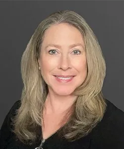 Smiling woman with long blonde hair wearing a black top against a dark background.