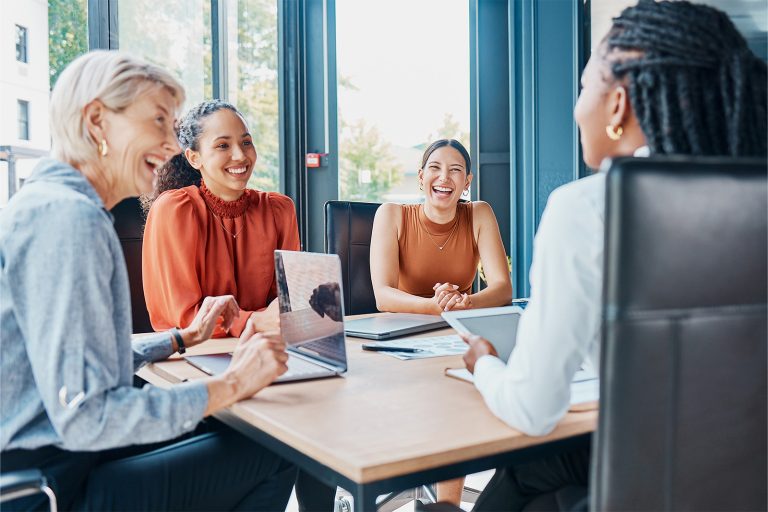 Four diverse women smiling and engaging in a lively meeting around a conference table with laptops and documents.