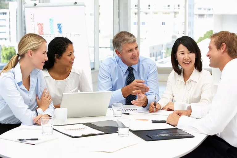 Five diverse business professionals seated around a conference table in a bright office, engaged in a discussion with documents, a laptop, and glasses of water visible.