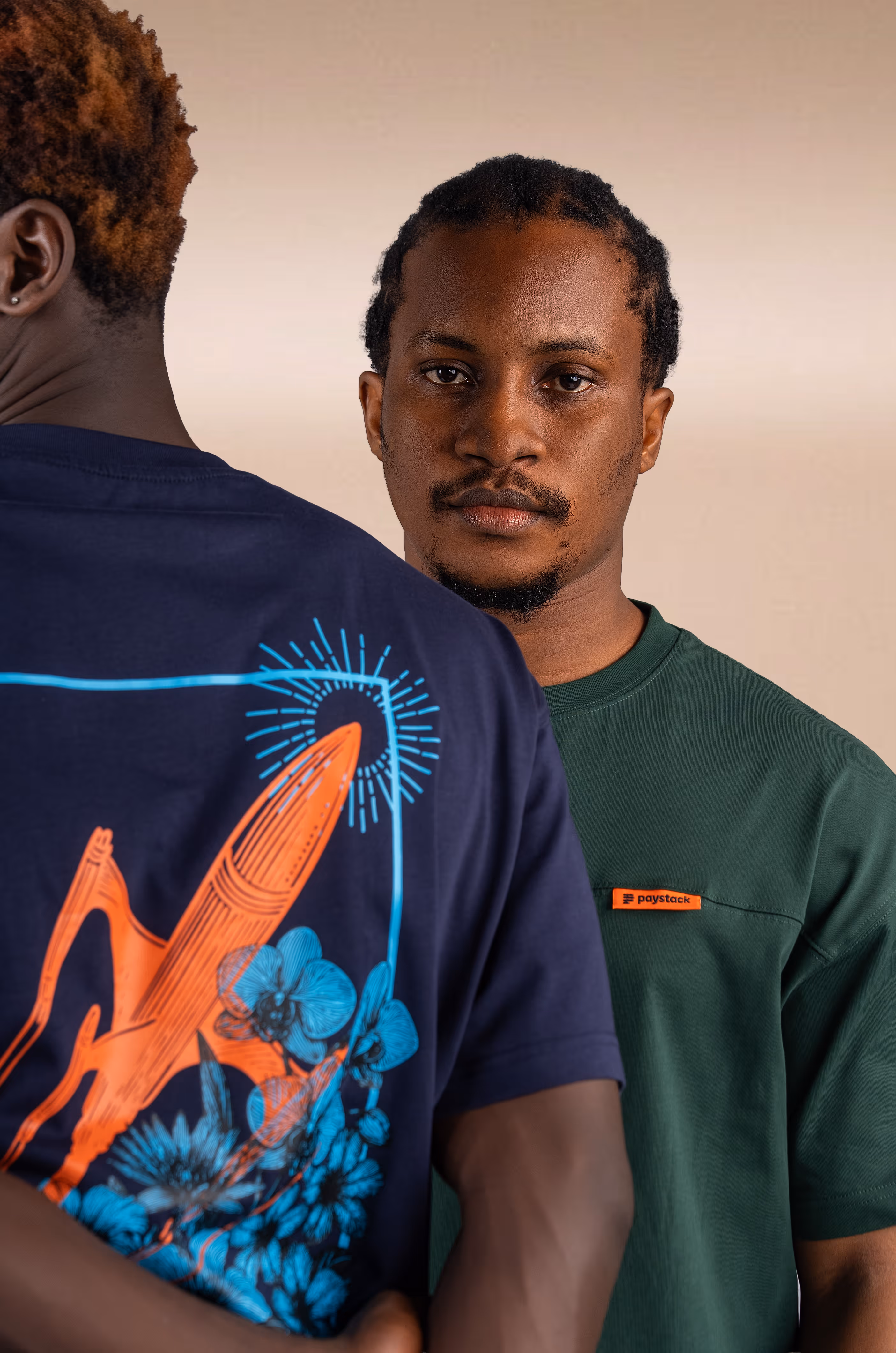 Two men standing closely; one faces the camera wearing a green shirt with an orange Paystack logo, the other turned away in a navy shirt with an orange rocket and blue flowers graphic.