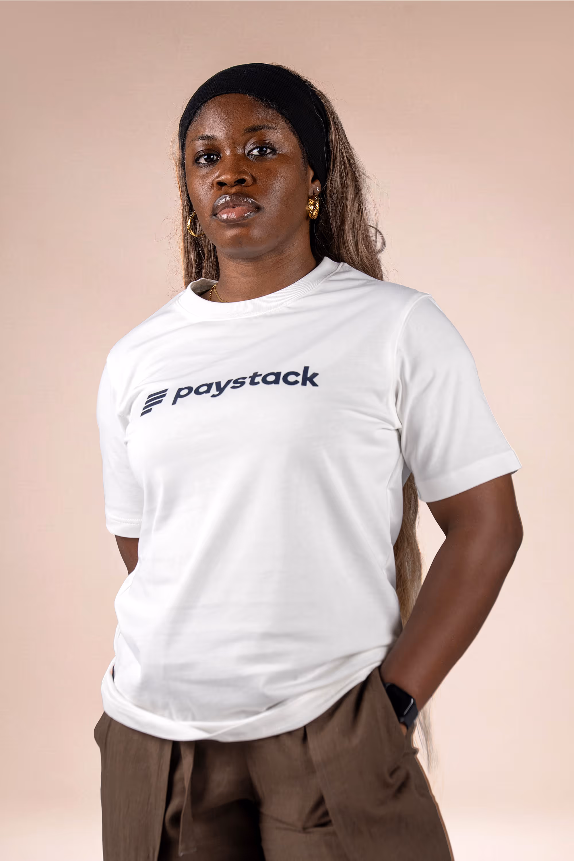 Woman wearing a white T-shirt with the word 'paystack' printed on it, brown pants, black headband, and gold hoop earrings.