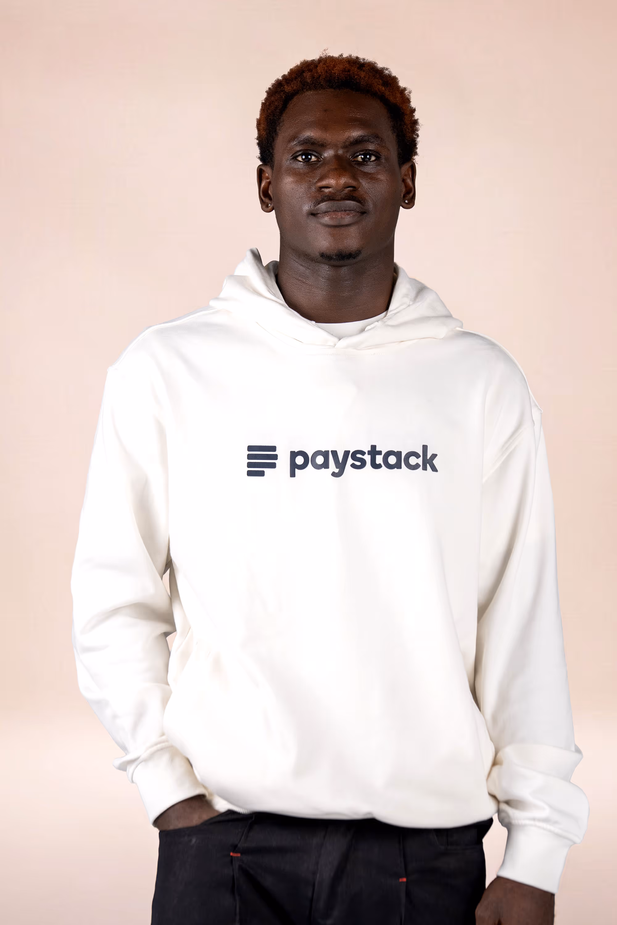 Man wearing a white hoodie with the Paystack logo on the front against a light pink background.