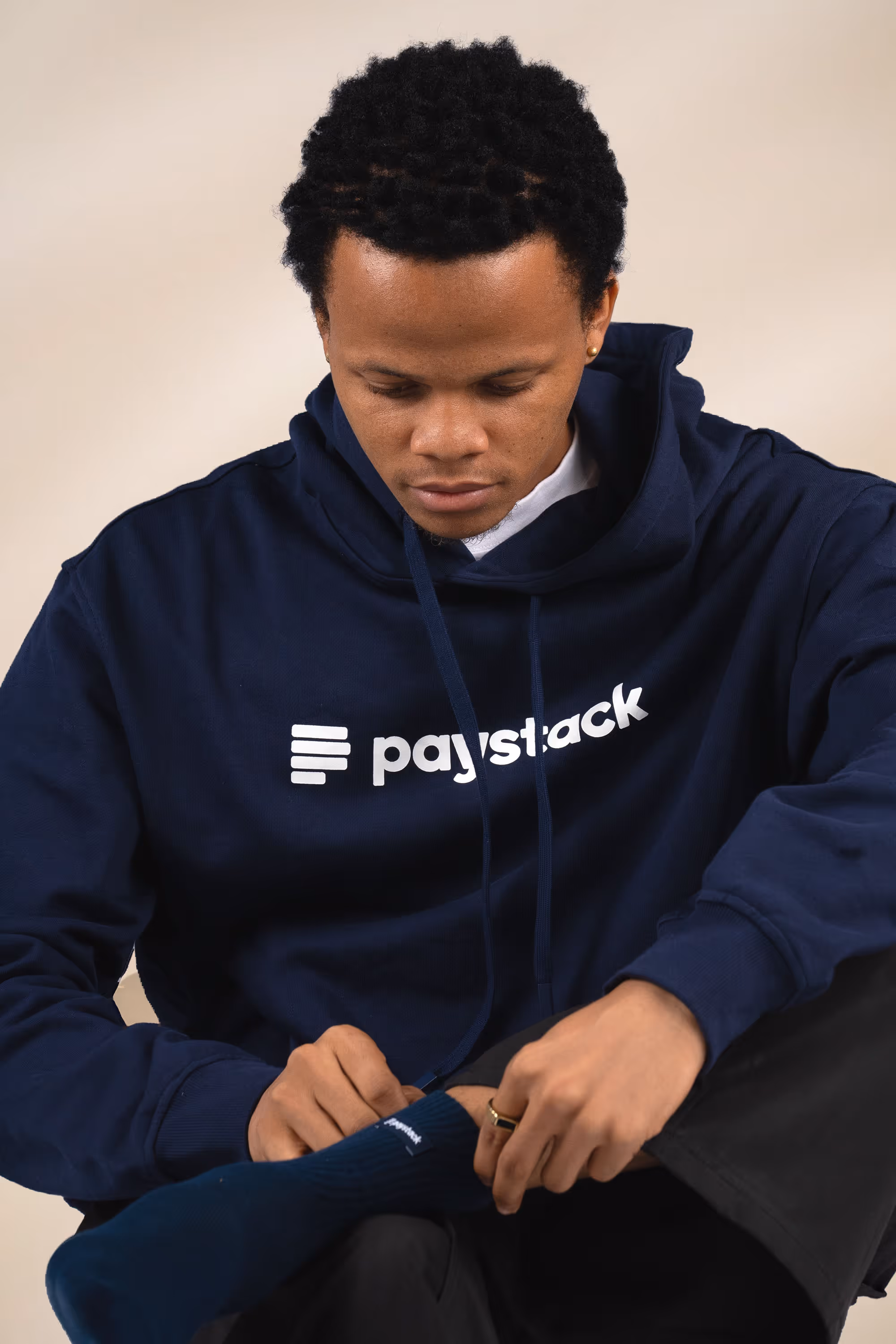 Man wearing a navy blue hoodie with the Paystack logo adjusting a navy sock while sitting with one leg crossed.