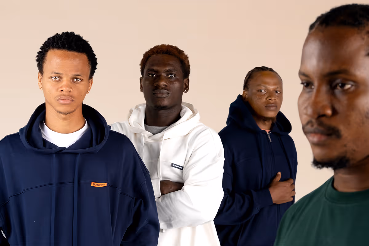 Group of four young men standing in a row against a beige background, wearing casual hoodies and shirts with serious expressions.
