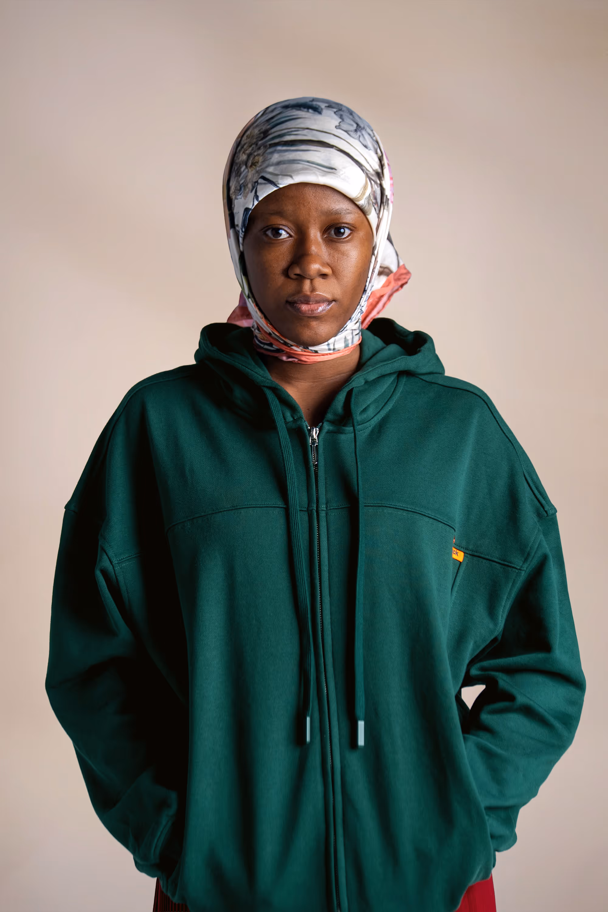 Woman wearing a dark green hoodie and a floral-patterned headscarf looking directly at the camera.