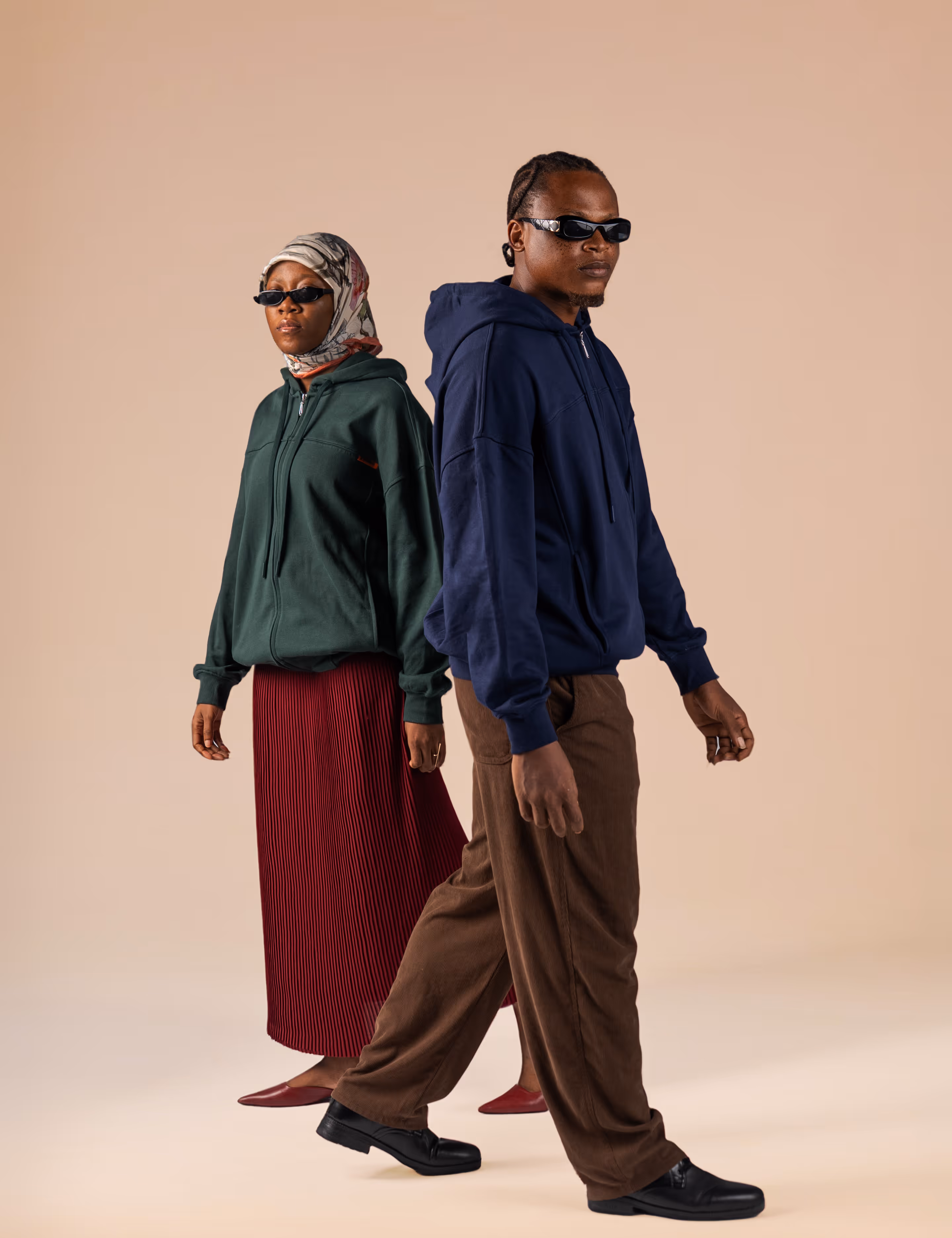 A man and woman wearing sunglasses and casual hoodies stand back to back against a beige background.