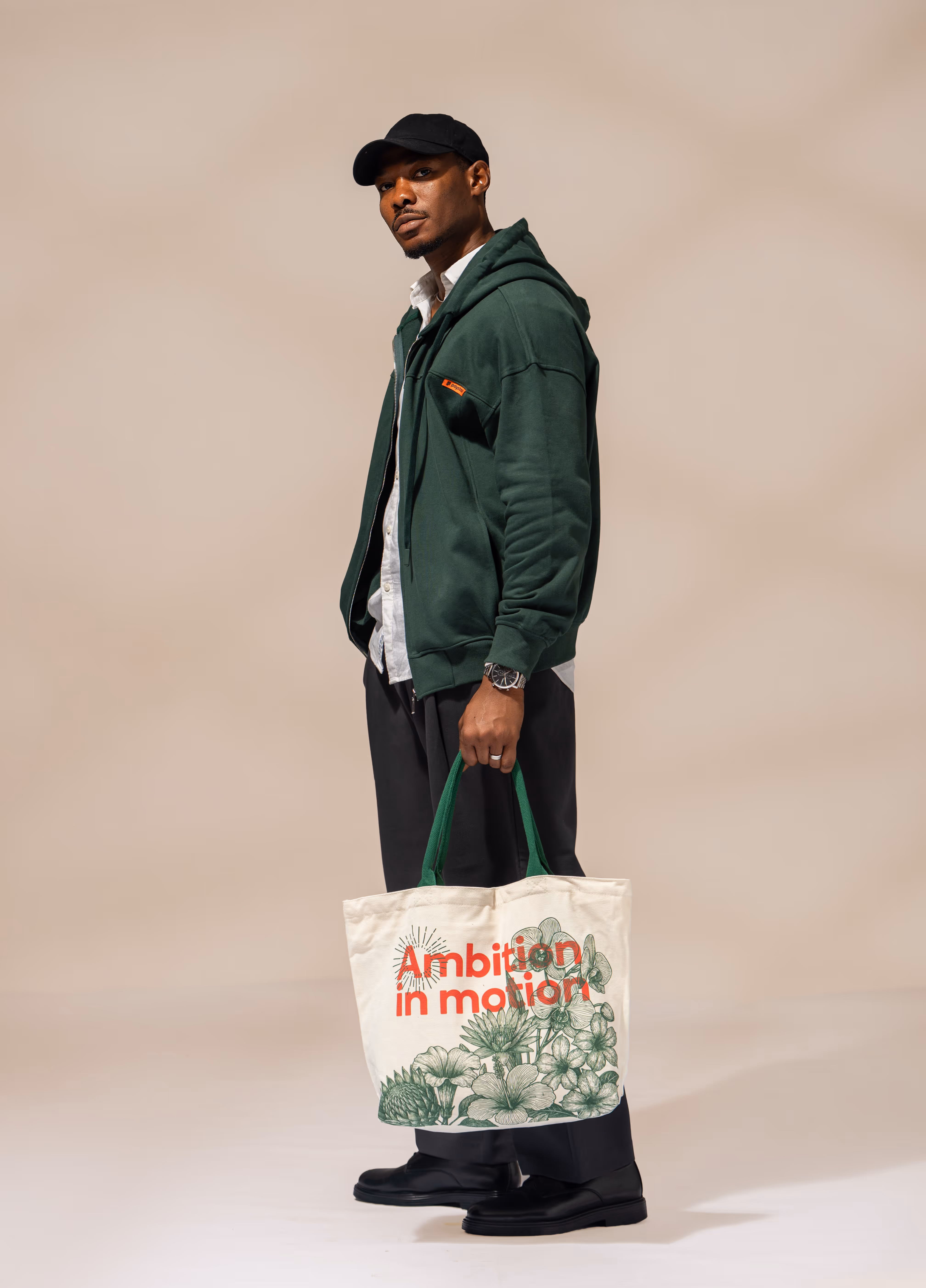 Man wearing a black cap, green hoodie, white shirt, and black pants holding a cream tote bag with green floral design and red text saying 'Ambition in motion'.
