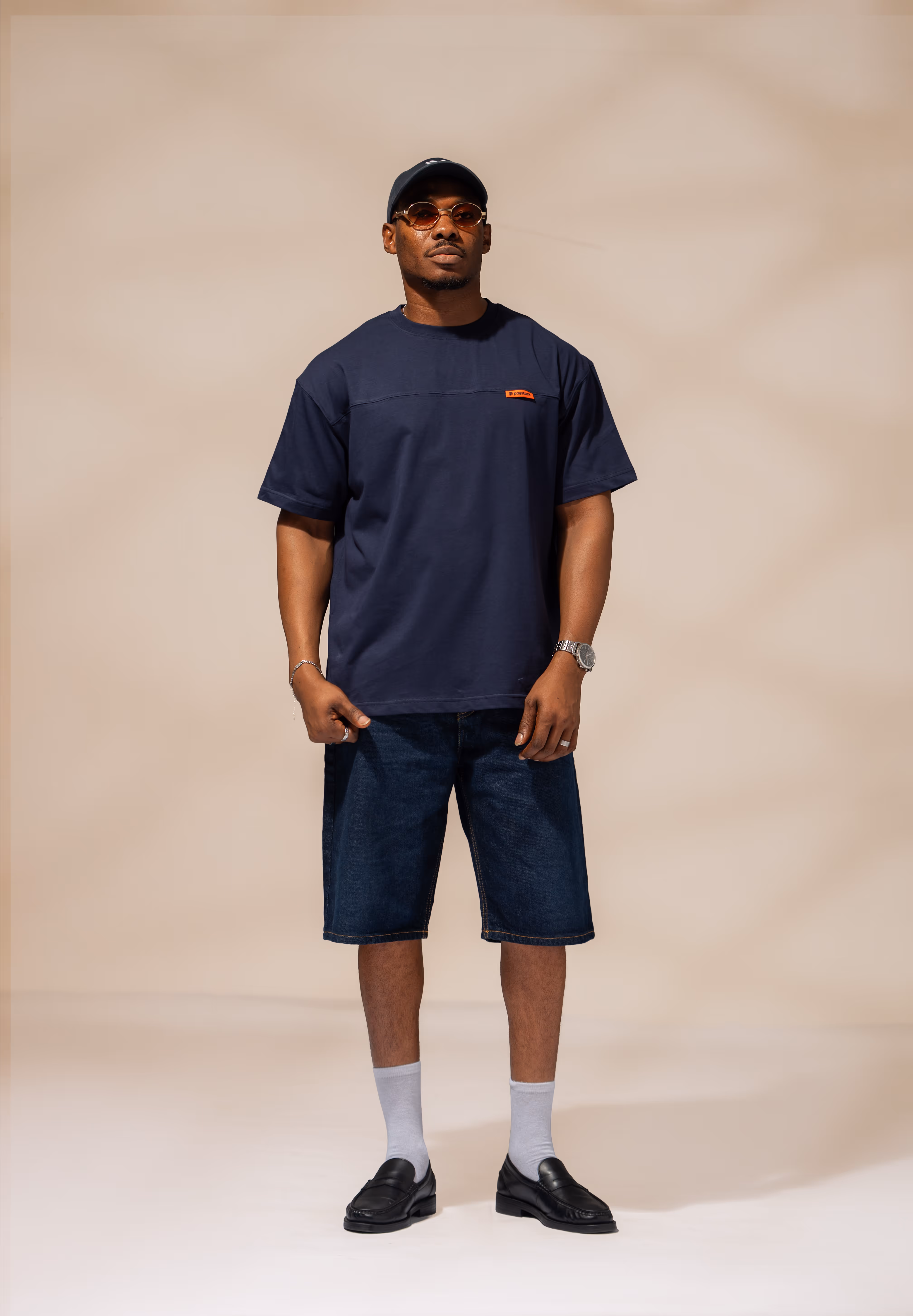 Man wearing a navy blue t-shirt, dark denim shorts, black loafers with white socks, a navy cap, brown sunglasses, and silver watch and bracelet, standing against a beige background.