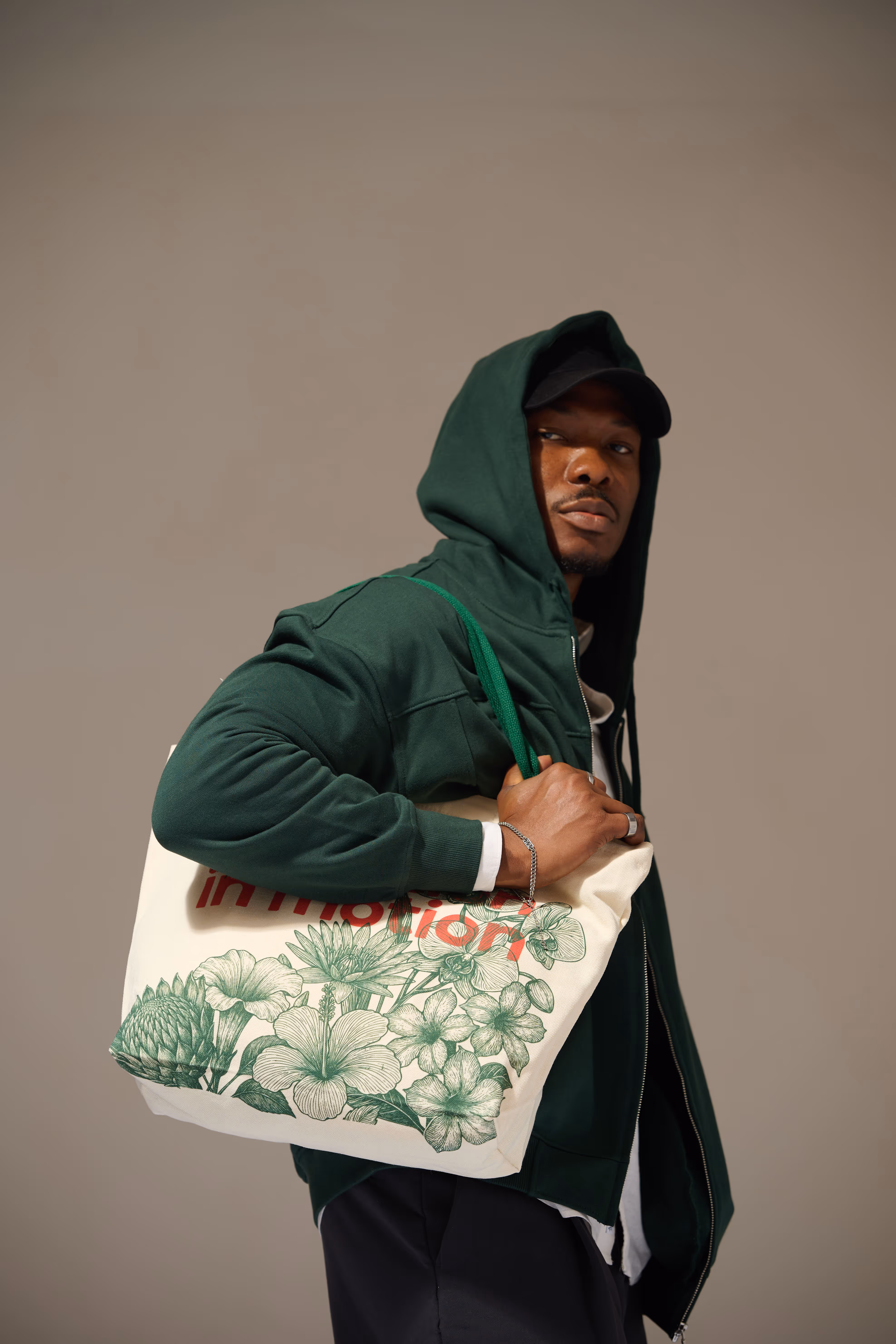 Man wearing a green hoodie and black cap carrying a beige tote bag with green floral print and red text.