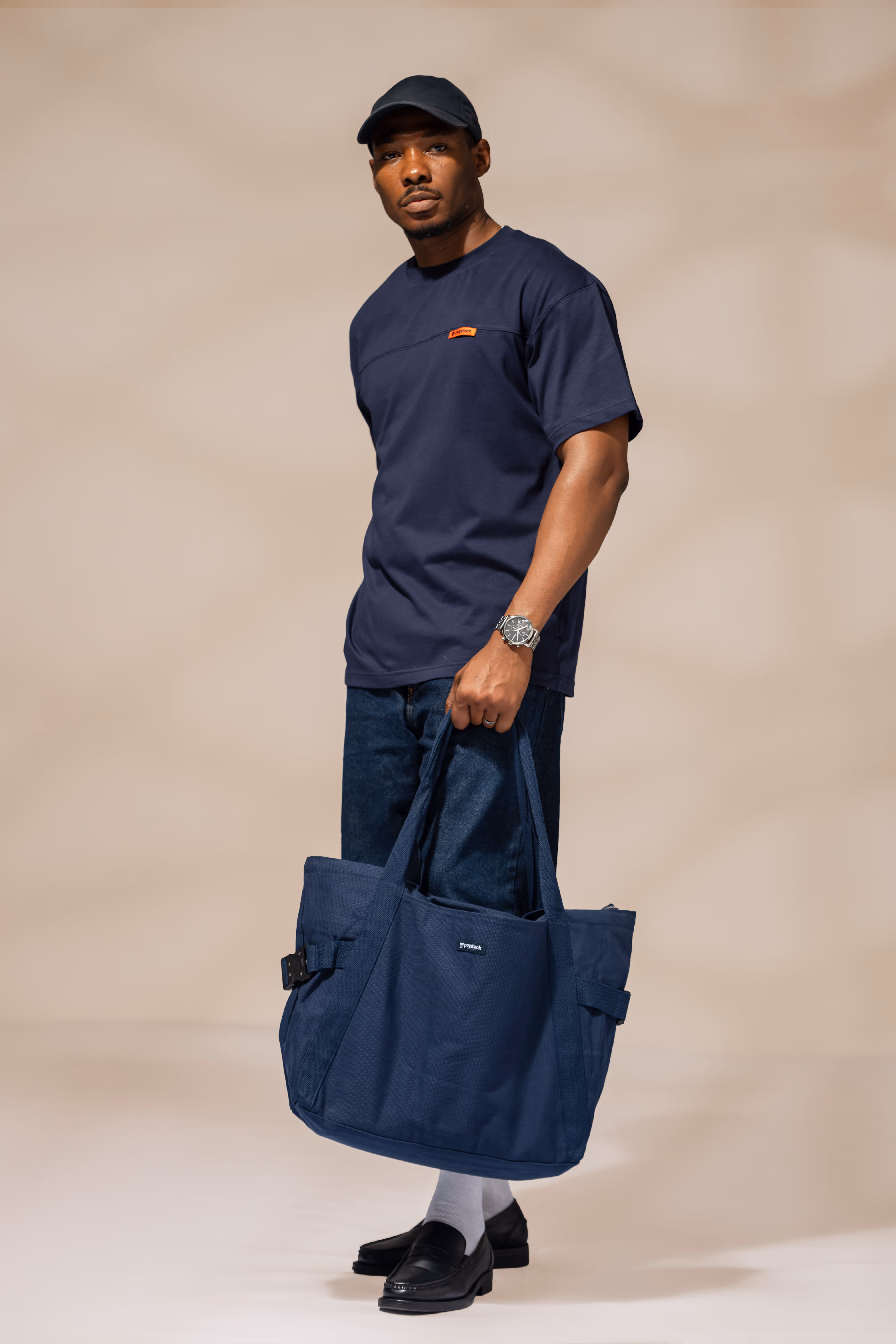 Man wearing a navy blue t-shirt, dark jeans, black cap, black loafers, and white socks holding a large navy blue tote bag.