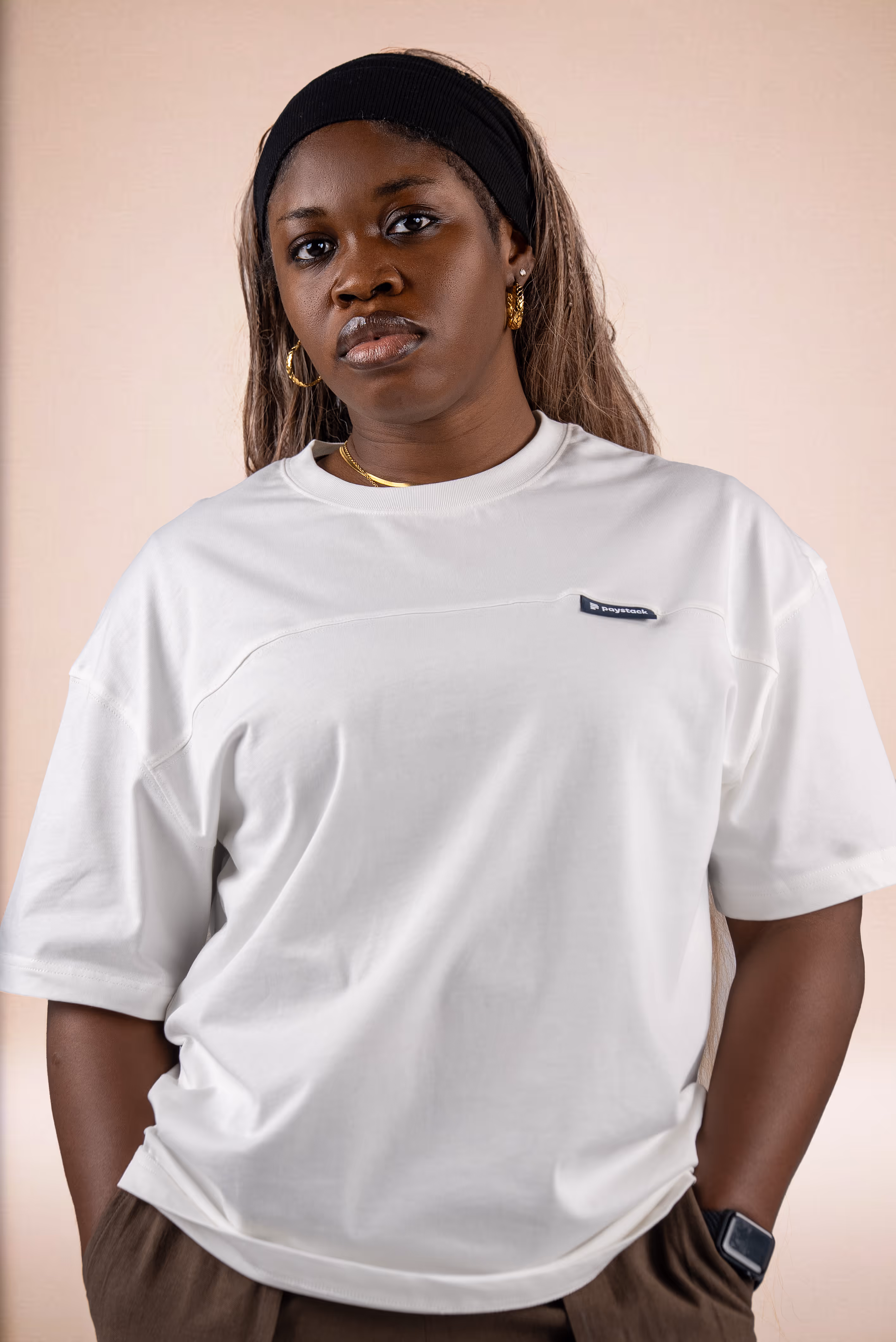 Portrait of a woman wearing a black headband, white t-shirt with a small logo, gold hoop earrings, and a smartwatch, against a plain beige background.