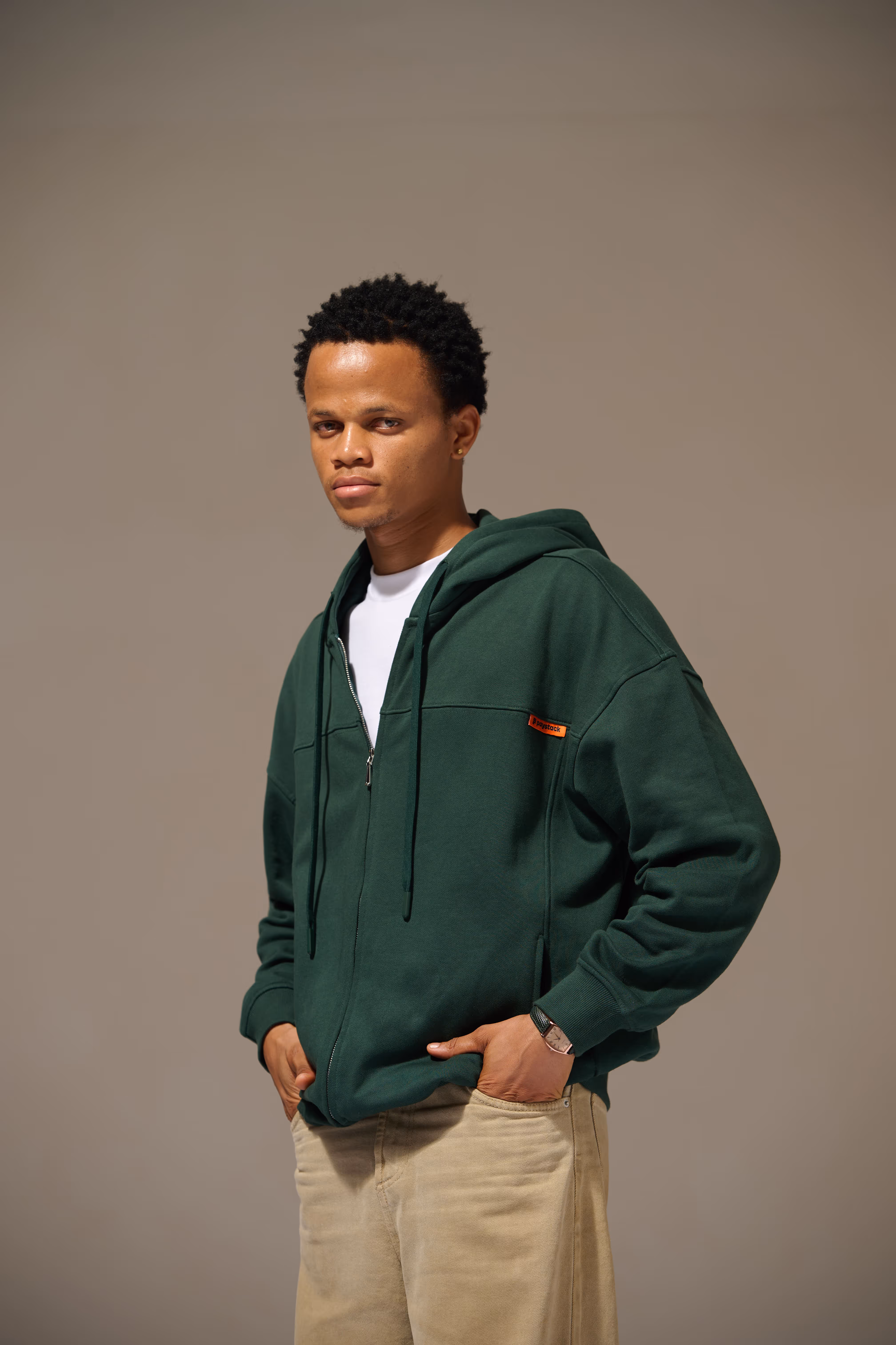 Man wearing a dark green zip-up hoodie and beige pants, standing with hands in pockets against a plain background.