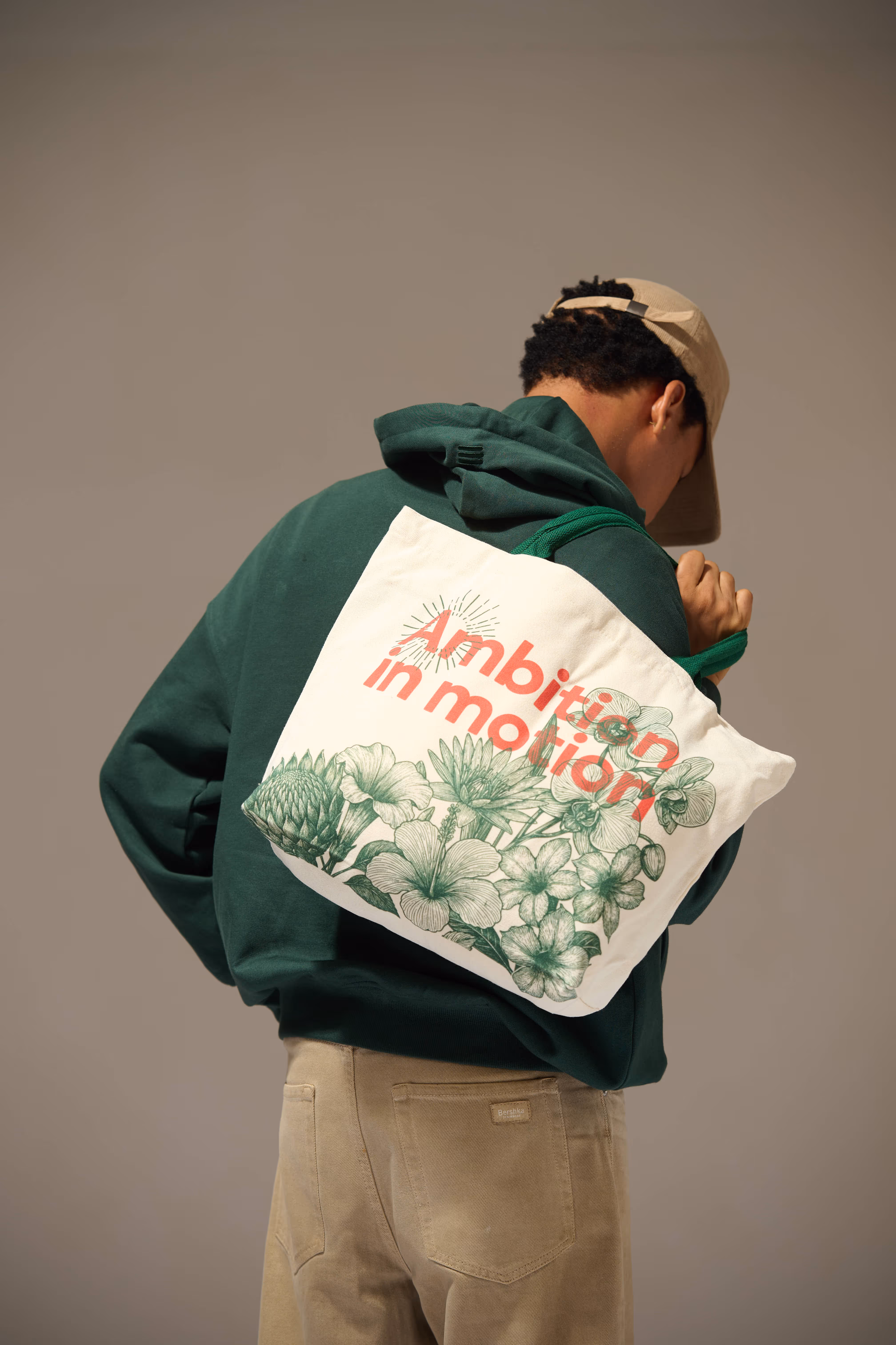 Person wearing a beige cap and green hoodie carrying a white tote bag with green floral designs and red text reading 'Ambition in motion'.