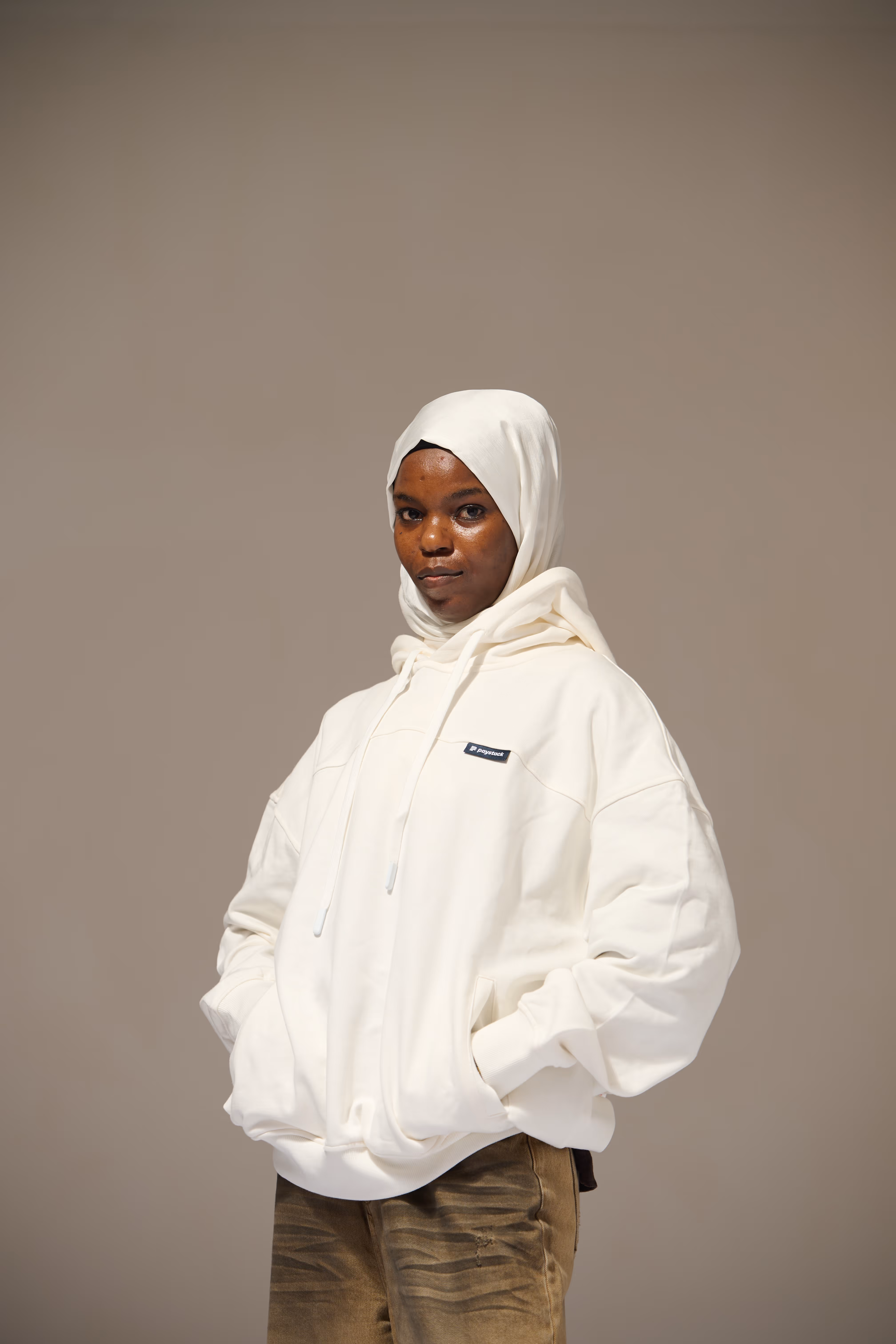 Person wearing a white hijab and oversized white hoodie with hands in pockets, standing against a plain background.