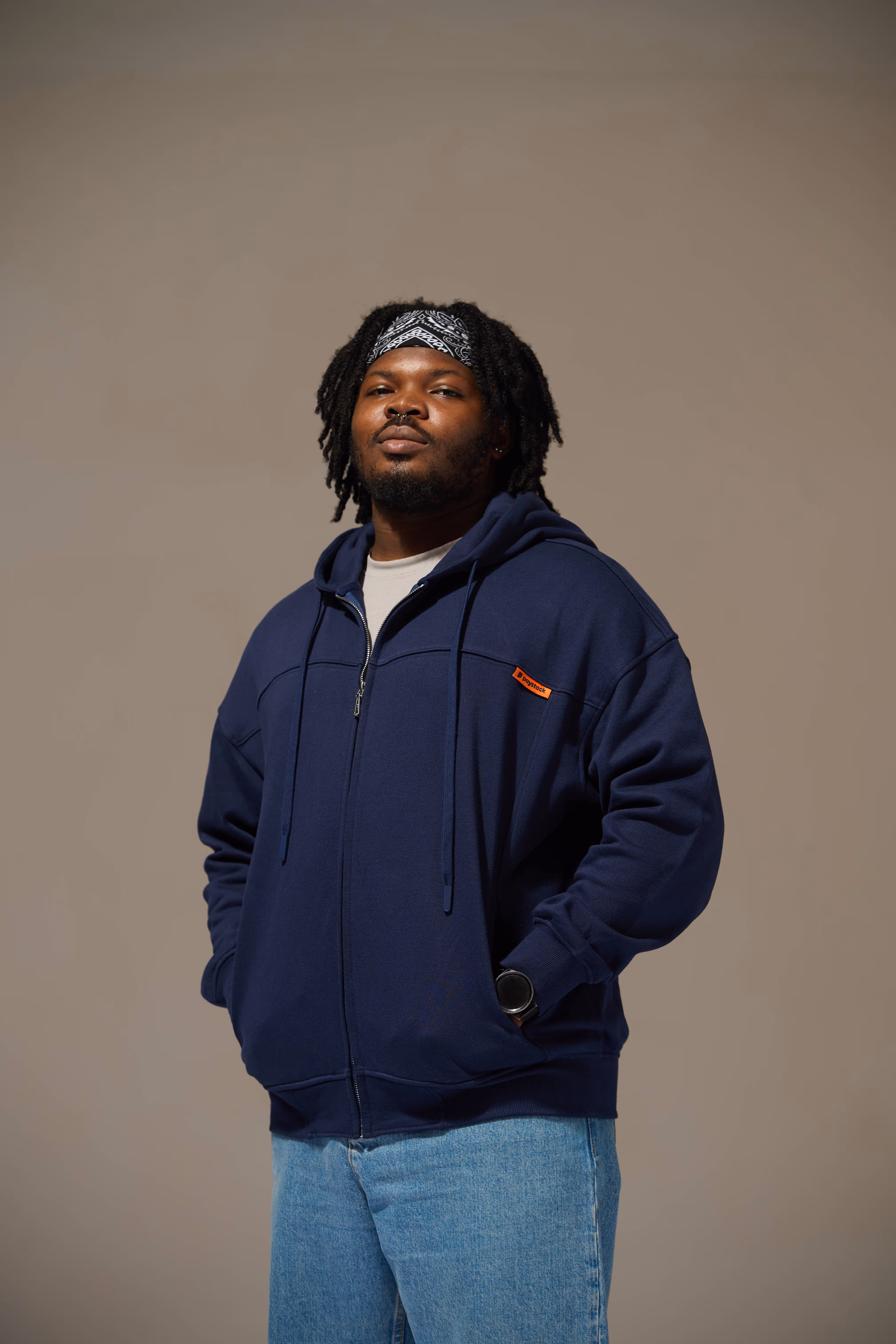 Man with dreadlocks wearing a navy blue hoodie, light blue jeans, and a black and white bandana standing against a plain background.