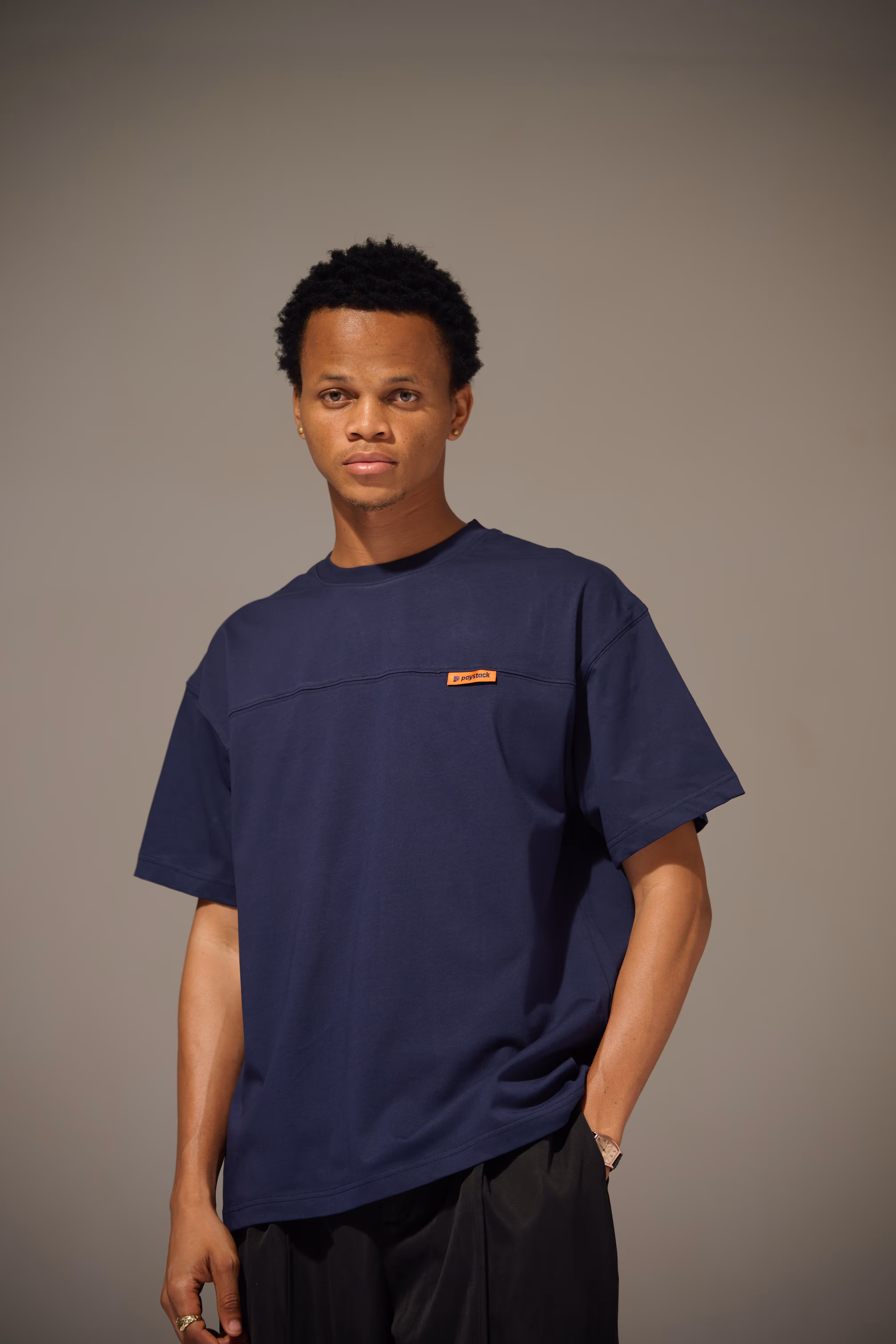 Young man with short curly hair wearing a navy blue T-shirt and black pants, standing against a plain gray background.
