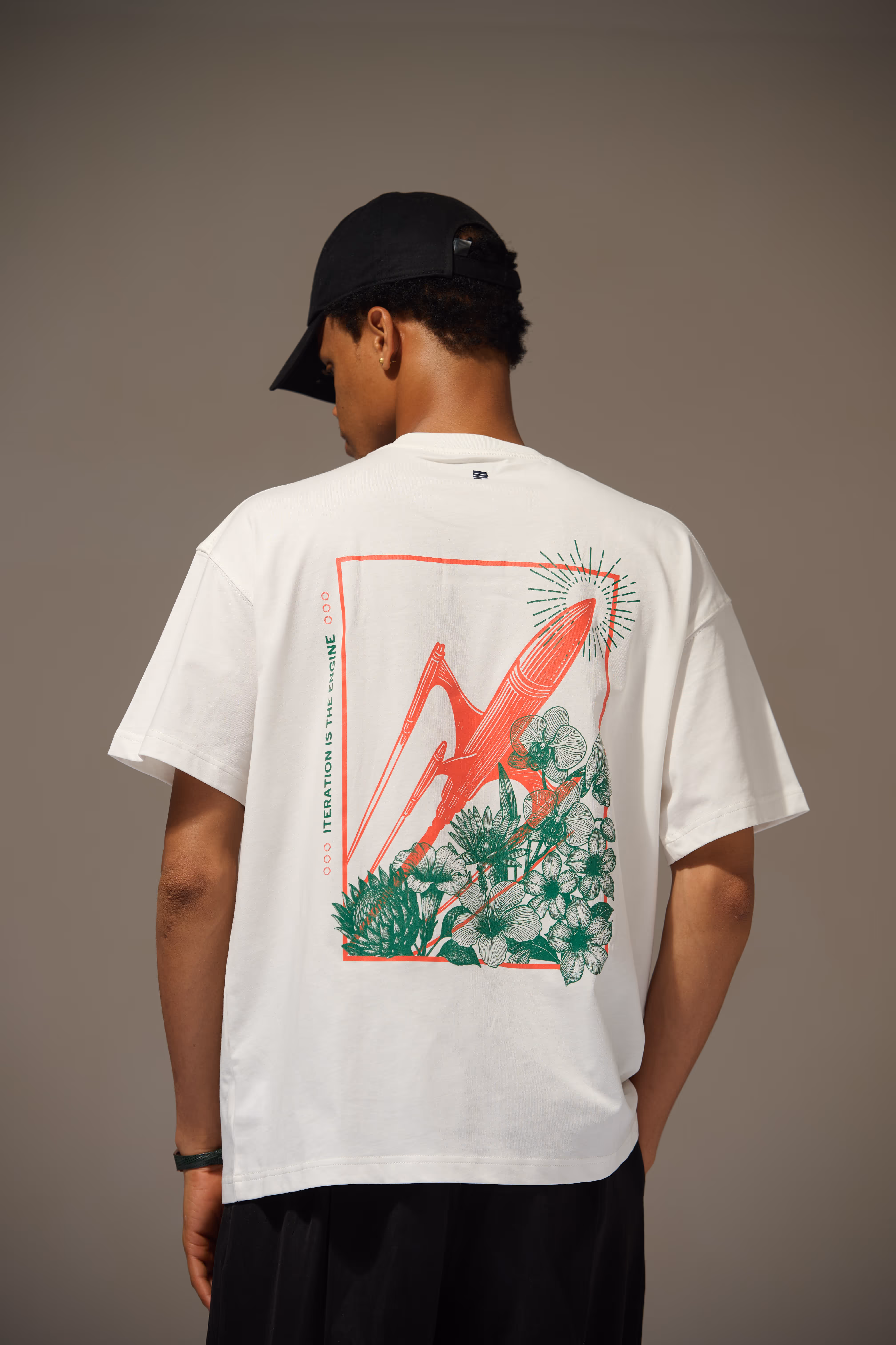 Man wearing a white T-shirt with a red and green graphic design of a rocket and plants on the back and a black cap.