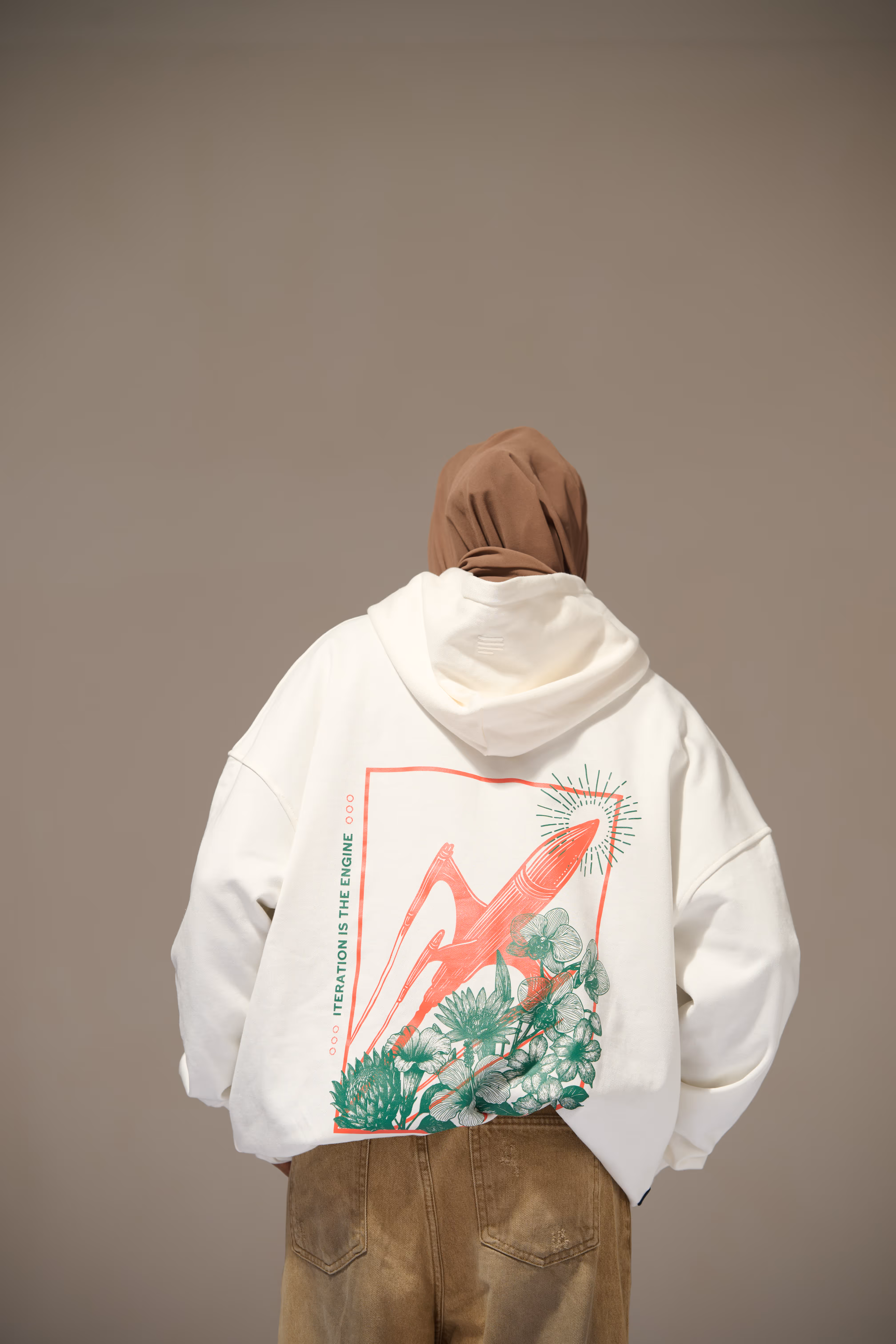 Person wearing a white hoodie with a red rocket and green floral design and the text 'ITERATION IS THE ENGINE' on the back, paired with brown pants and a brown headscarf.