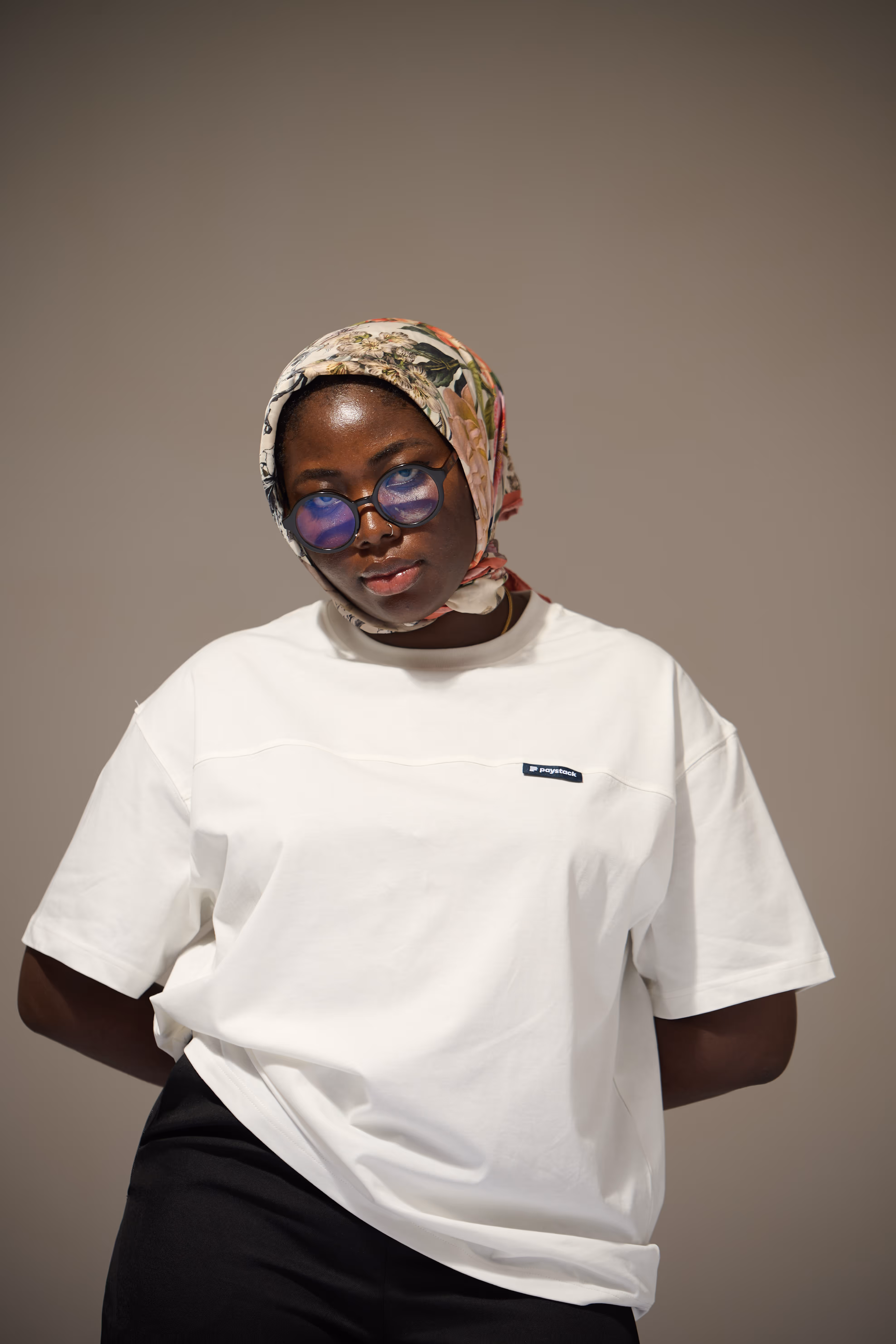 Person wearing a floral headscarf, blue-tinted round glasses, and a white t-shirt with a small logo on the chest.