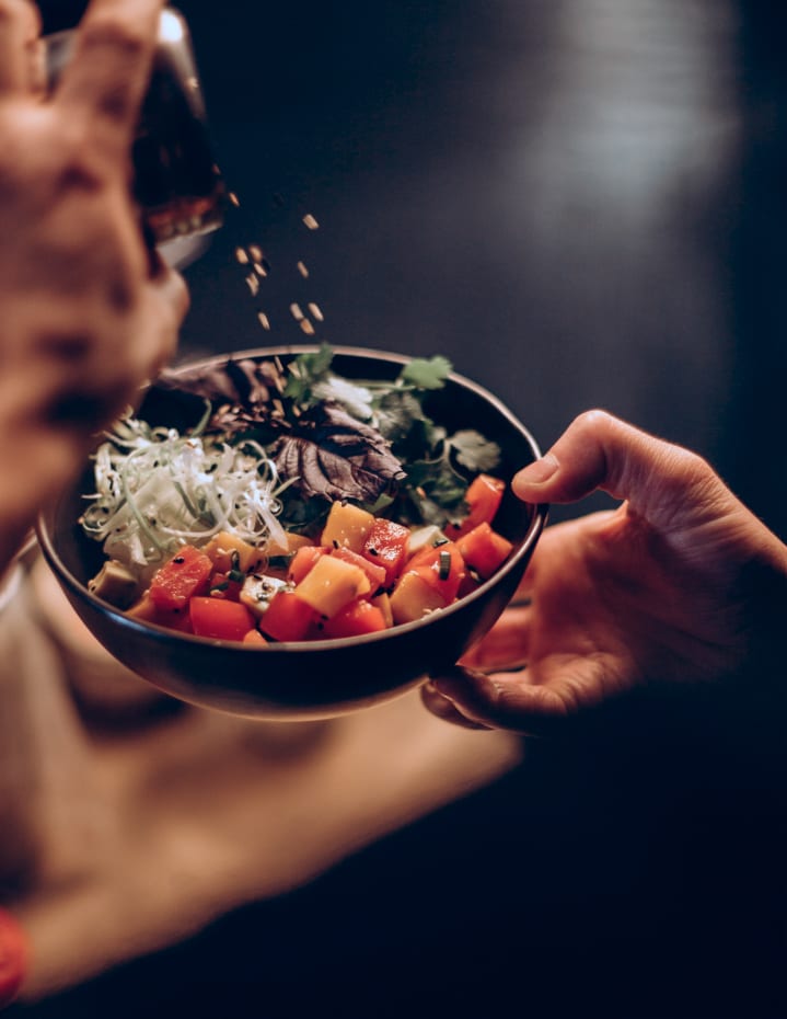 Une main tenant un bol de salade colorée pendant qu'une autre main saupoudre des graines dessus.