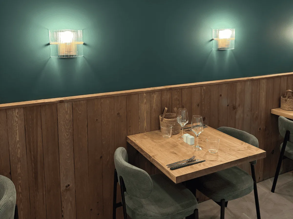 Coin repas avec une table en bois clair dressée pour deux personnes, deux chaises vert mousse et un mur avec lambris en bois et luminaires muraux modernes.