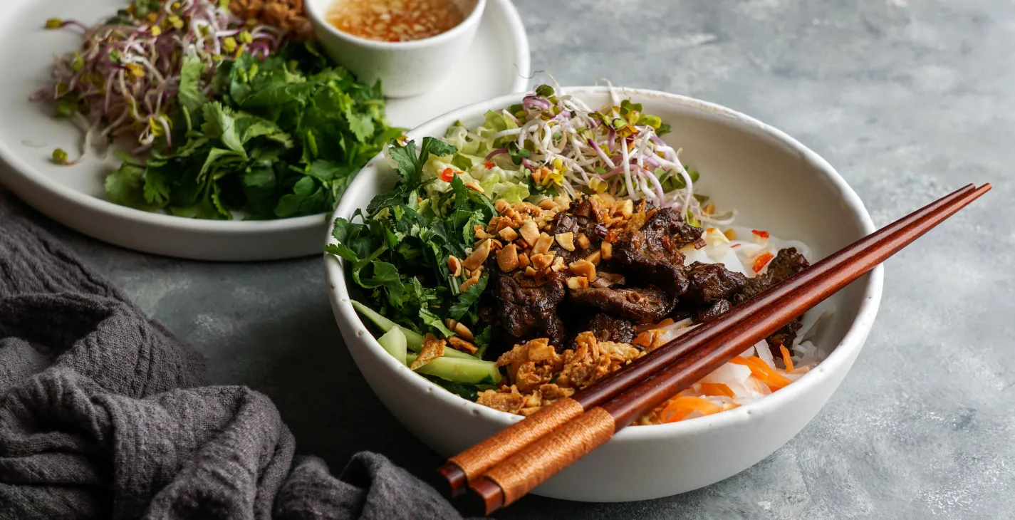 Bol vietnamien avec bœuf mariné, légumes frais, herbes, cacahuètes concassées, germes, et baguettes en bois posées dessus à meicha cysoing