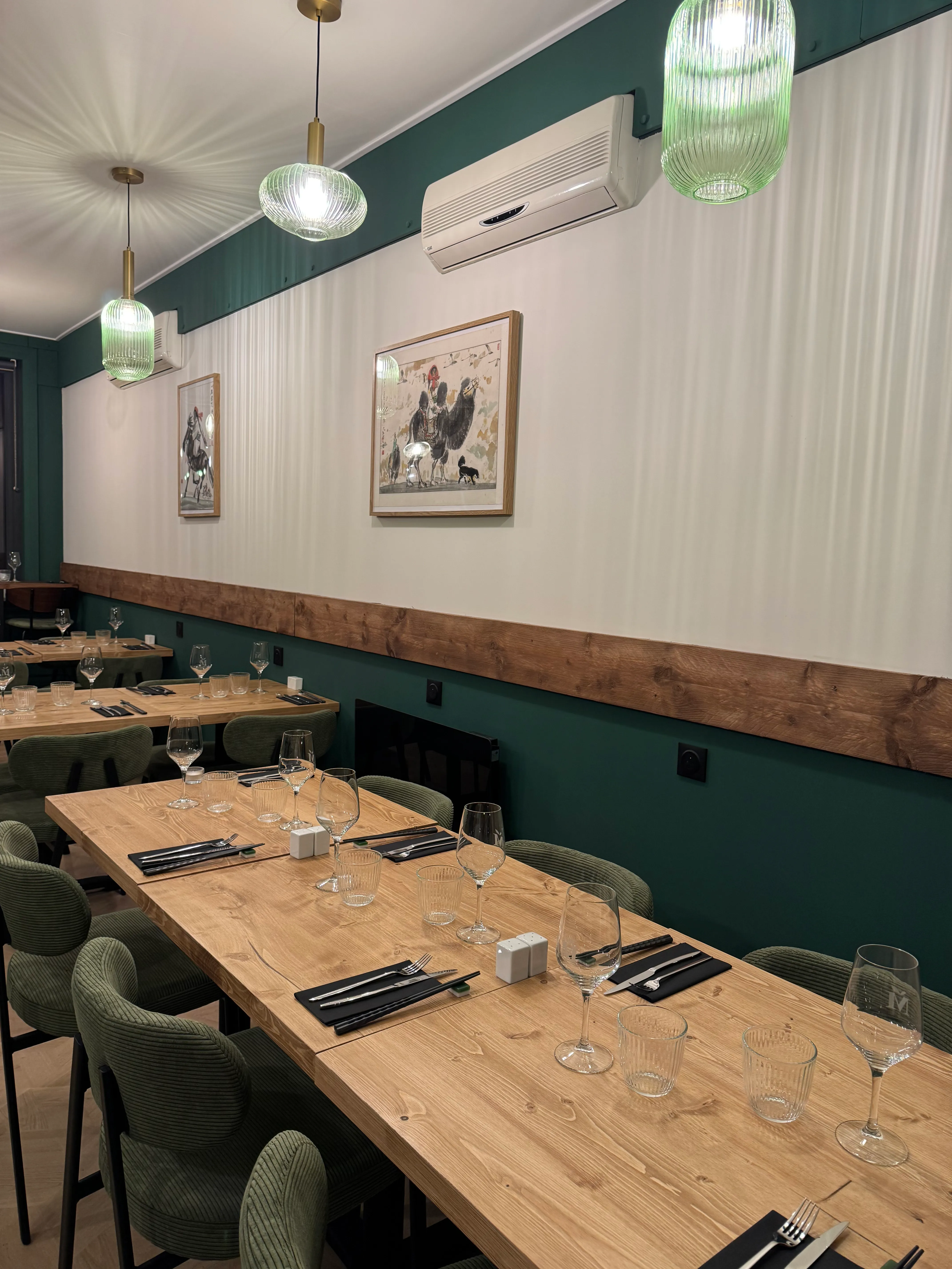 Intérieur d'un restaurant avec une grande table en bois, des verres à vin, des couverts sur des serviettes noires, des chaises vertes, des suspensions lumineuses en verre et deux cadres de peinture accrochés au mur meicha