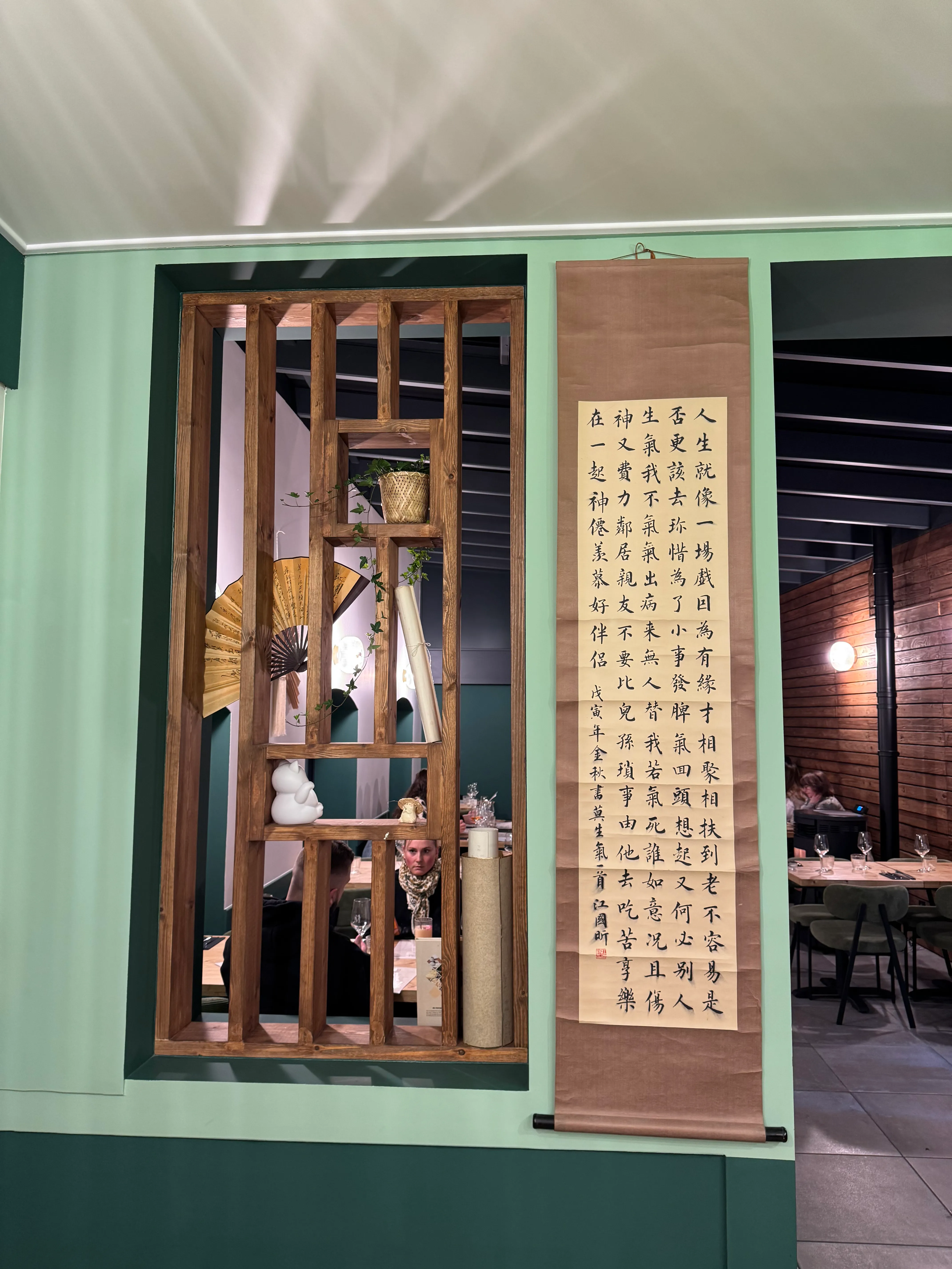 Intérieur de restaurant avec une cloison en bois décorative, une calligraphie chinoise accrochée au mur et des clients assis autour d'une table à Meicha