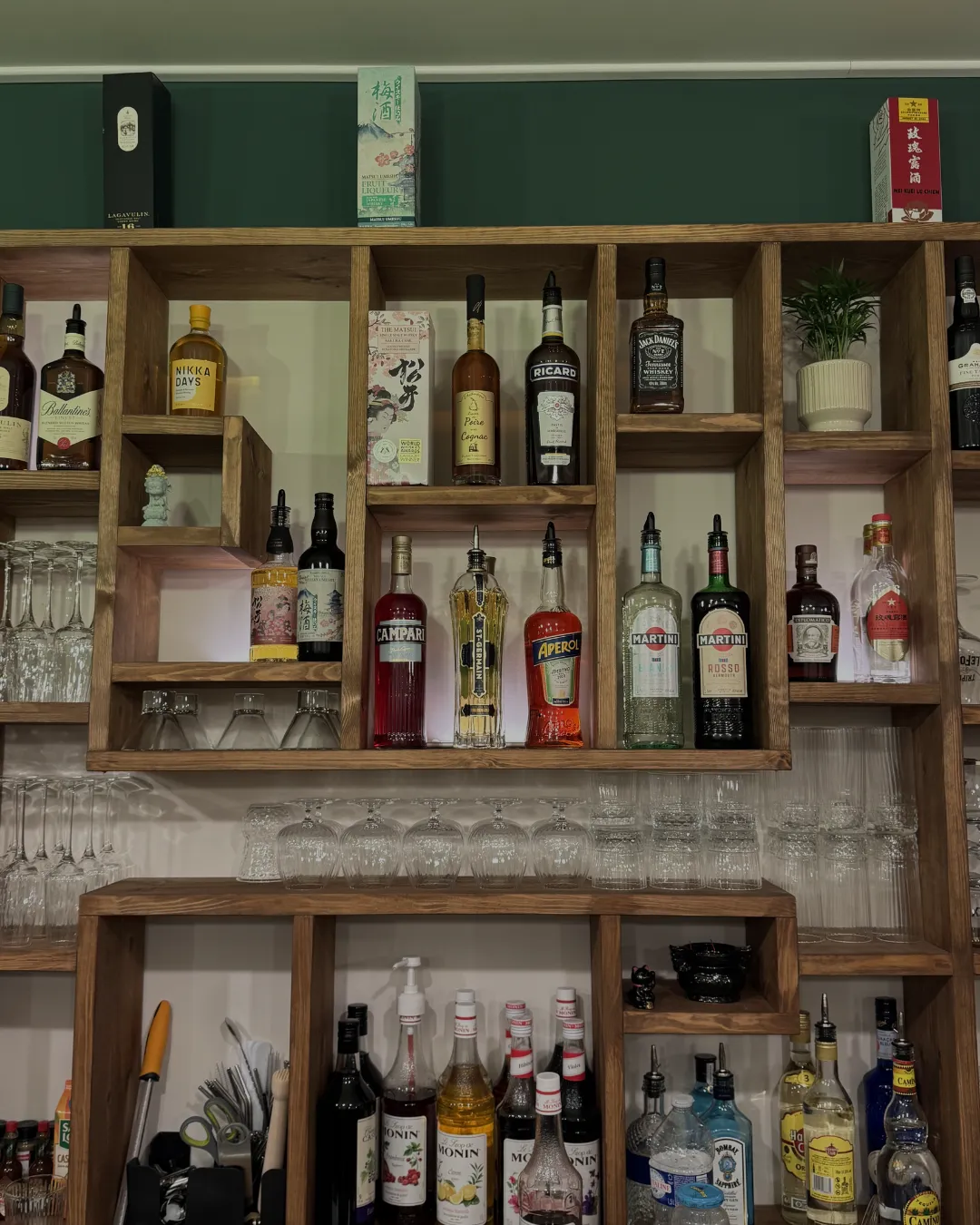 Étagère en bois avec diverses bouteilles d’alcool, verres à pied, et un pot de plante, organisés dans des niches cubiques.