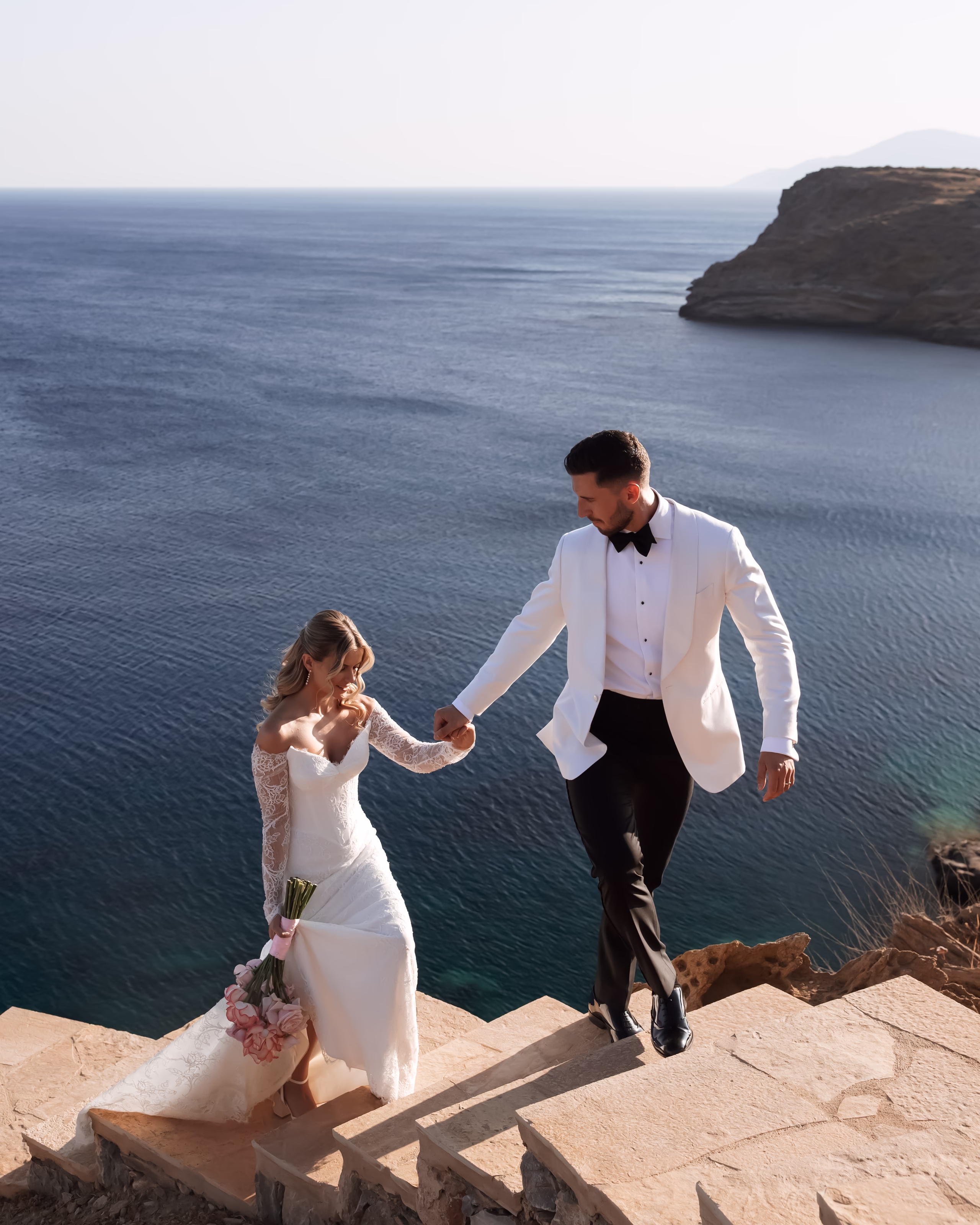 A bride in a lace gown and a groom in a white tuxedo jacket holding hands while walking down stone steps by the sea.