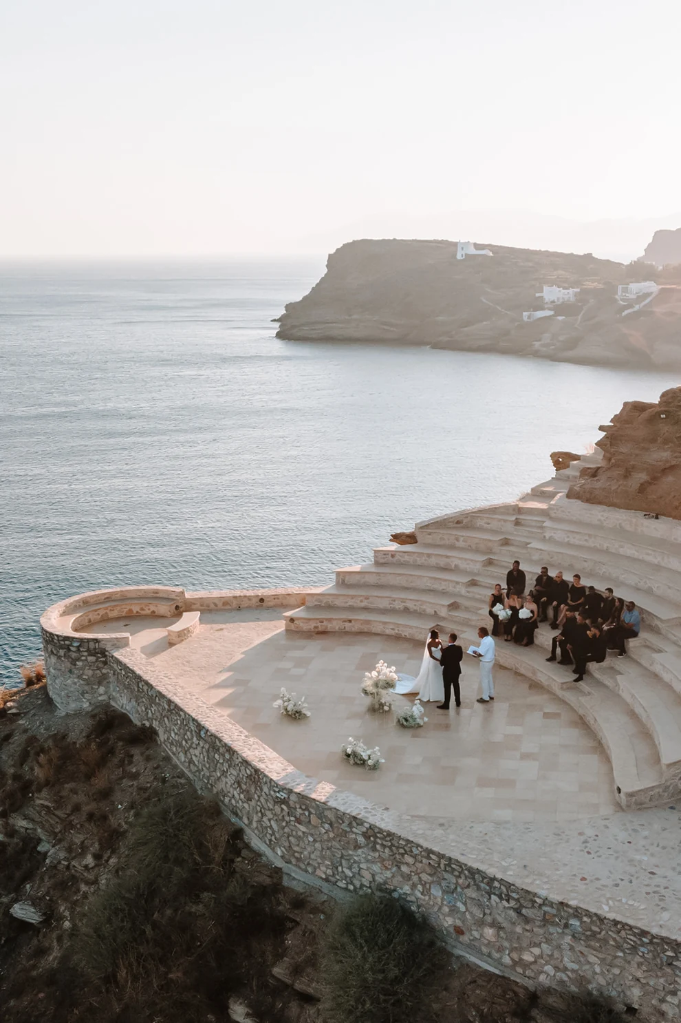 An aerial view of an intimate wedding ceremony held in a stone amphitheater overlooking a dramatic Mediterranean coastal vista.