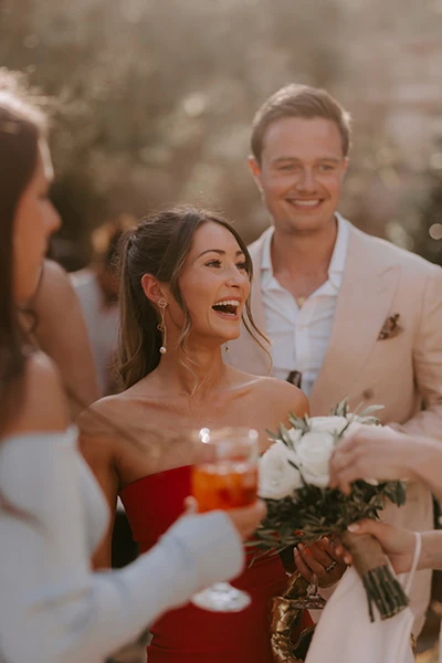 Smiling woman in red dress laughing while holding white roses and a drink at a social gathering.