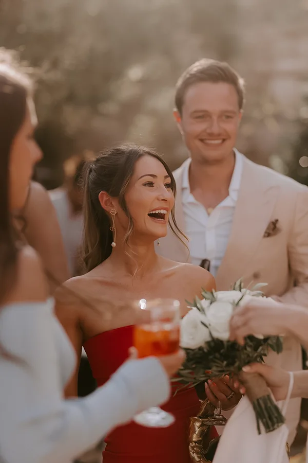 Smiling woman in red dress laughing while holding white roses and a drink at a social gathering.