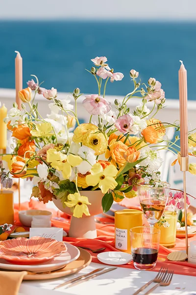 Table set with colorful floral centerpiece, candles, glassware, and plates overlooking a blue ocean background.