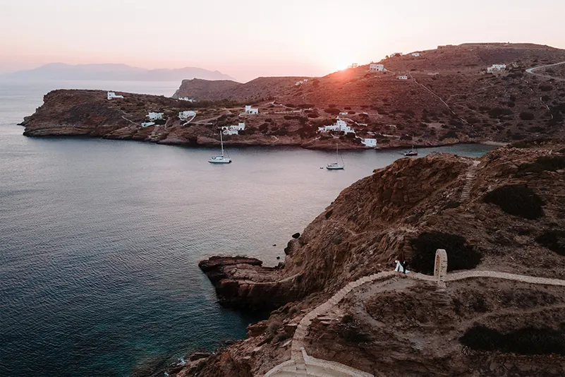 A breathtaking sunset over a bay in Ios, Greece, with sailboats anchored near the rocky coastline and white coastal dwellings.