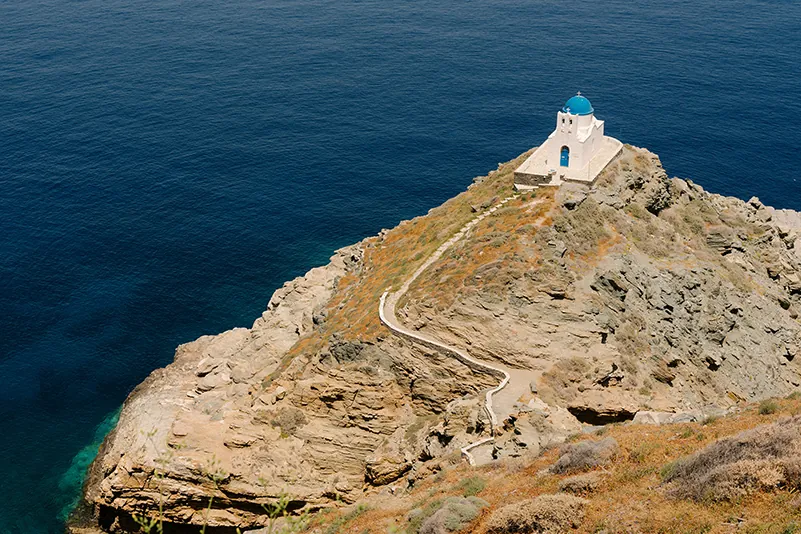 The iconic white church with a blue dome in Sifnos, perched on a rocky peninsula surrounded by deep blue waters.